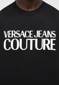 Μαύρο μπλουζάκι με μεγάλο λευκό κείμενο που γράφει "VERSACE JEANS COUTURE" στο στήθος, φορεμένο από ένα άτομο.
