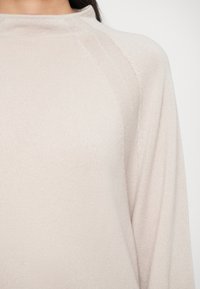 Maglione beige chiaro con texture a coste, colletto alto e maniche a raglan. Tessuto morbido, messo in risalto da sottili dettagli di cucitura.