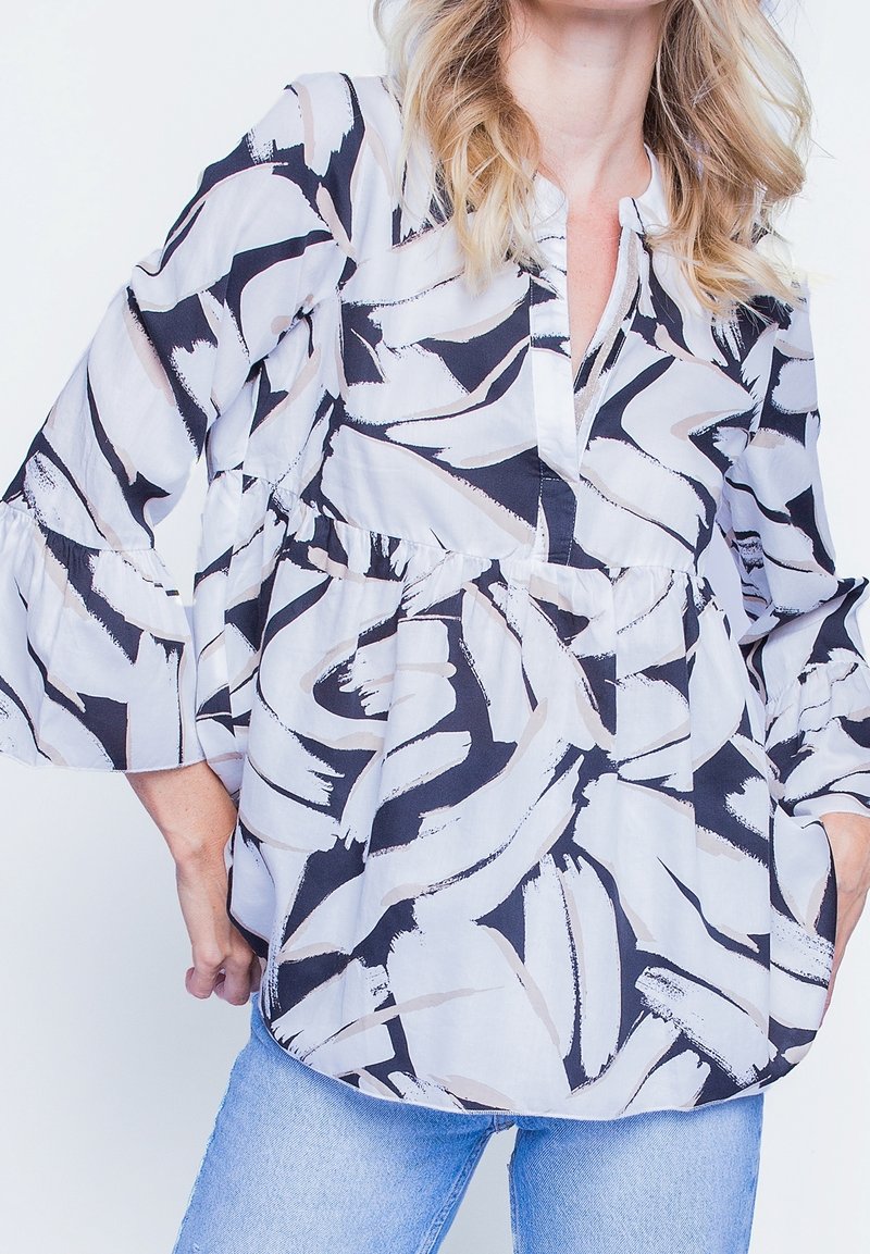 Femme portant un chemisier ample à manches longues avec des motifs abstraits noirs et blancs et un jean bleu clair sur fond blanc.