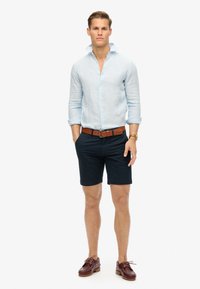 Lichtblauw linnen shirt met opgerolde mouwen, bruine leren riem, marineblauwe shorts en bruine bootschoenen. Zachte textuur, casual pasvorm.