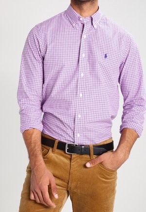 Homme portant une chemise à carreaux violette boutonnée rentrée dans un pantalon en velours côtelé beige avec une ceinture noire, une main dans la poche.