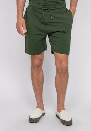 Homme portant un short vert foncé à cordon et des chaussures slip-on écrues, debout les mains dans les poches devant un fond uni.