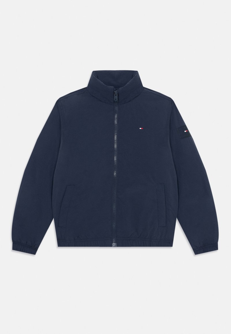 Veste légère bleu marine avec un col haut, fermeture éclair à l'avant et deux poches latérales. Présente des poignets élastiques et un petit logo sur la poitrine.