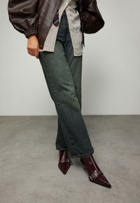 Bruine leren bomberjack met opgeblazen mouwen, gestreept overhemd, donkergroene wijde spijkerbroek en bordeauxrode puntige laarzen met zilveren accenten.