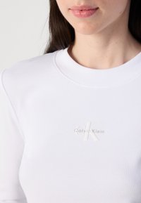 Un primo piano della parte superiore del corpo di una donna che indossa un top bianco a coste con un sottile logo Calvin Klein sul petto, mettendo in risalto la pelle liscia e le labbra.