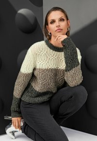 Frau kniet und trägt einen grob gestrickten gestreiften Pullover in Cremetönen, Braun und Dunkelgrün, schwarze Hosen, silberne Ohrringe und metallic-silberne Stiefel.