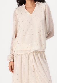 Blouse beige avec manches longues et transparentes et encolure à volants ; ornée de pois métalliques ; ourlet en couches ajoutant de la texture.