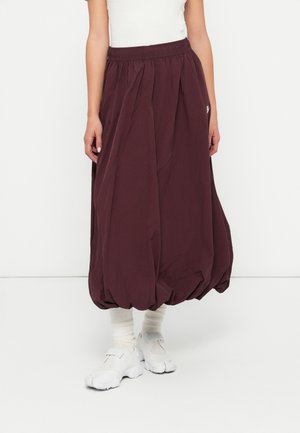 Person iført en mørkelilla ballon-nederdel, hvide sokker og chunky hvide sneakers, der står mod en ensfarvet hvid baggrund.