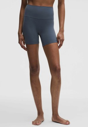 Person steht barfuß und trägt eng anliegende, dunkelgraue Sportshorts mit hoher Taille vor einem einfarbigen weißen Hintergrund.