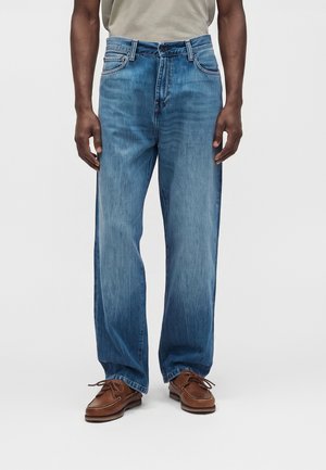Homme portant un jean bleu large et des chaussures en cuir marron à lacets, debout devant un fond clair uni.