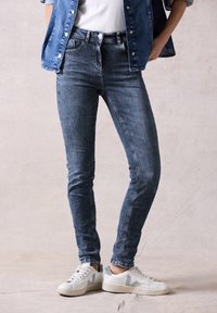 Jeans ajustados en denim azul desgastado, con una cintura de altura media, diseño de cinco bolsillos y un ligero efecto de desvanecimiento. Combinados con zapatillas blancas.