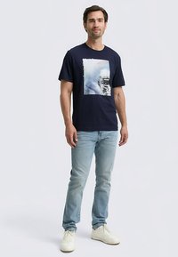 Tumši zils kokvilnas T-krekls ar grafisku attēlu ar transportlīdzekli, kombinēts ar gaišām džinsa biksēm un baltiem sporta apaviem. Ikdienišķs piegriezums.