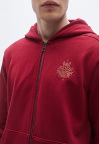 Červená mikina na zips z mäkkého materiálu, s pozláteným vyrazeným logom "BOSS" na hrudi a kapucňou so šnúrkami.