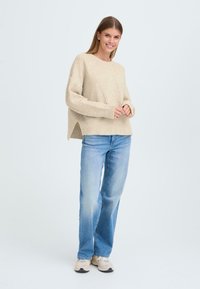 Beiger Strickpullover mit diagonaler Rippstruktur, seitlichen Schlitzen und lockerer Passform, kombiniert mit hellblauen Gerade-Leg-Jeans und beigen Sneakers.