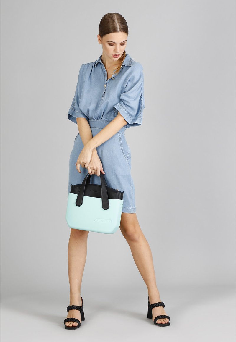 Borsa verde menta con manici neri, texture liscia e forma arrotondata, abbinata a un vestito in denim chiaro e sandali con tacco neri.