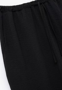 Zwarte loungebroek van zacht, lichtgewicht stof. Voorzien van een elastische tailleband met een trekkoord en zijzakken. Gladde textuur.