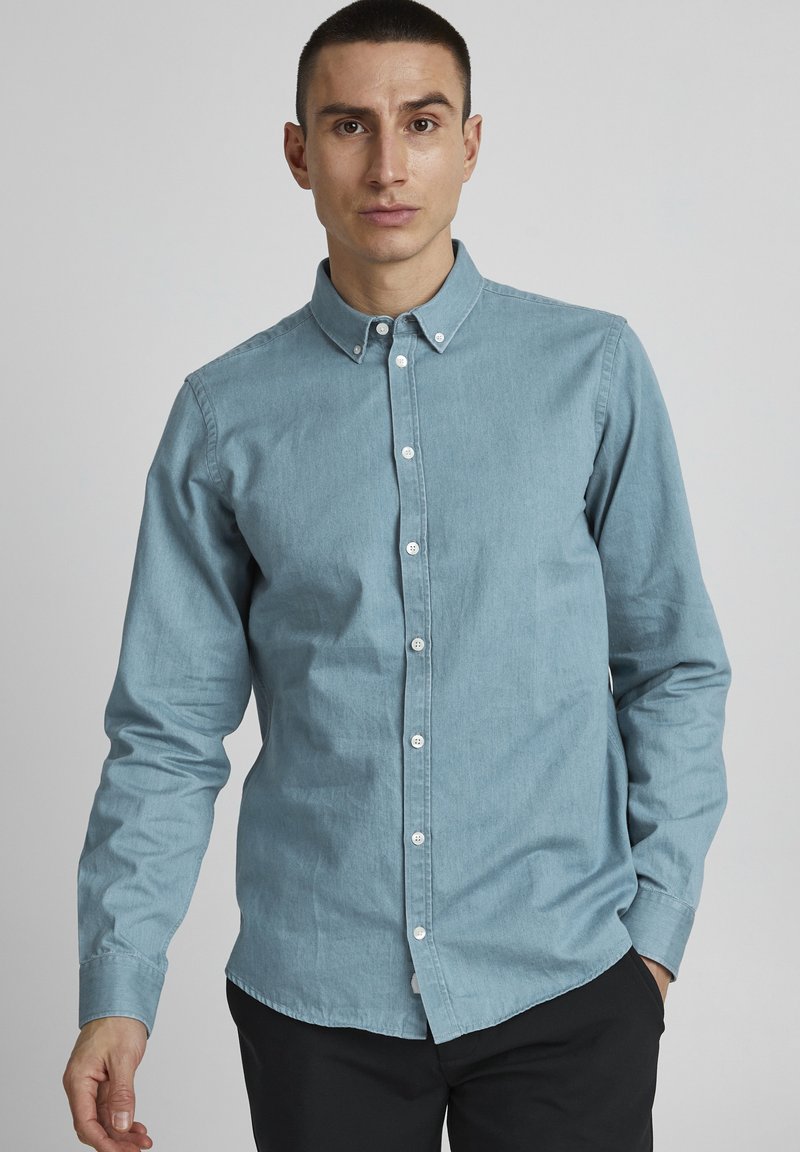 Light blue button-up shirt made of cotton fabric, featuring a classic collar, long sleeves, and a curved hem, paired with dark pants.