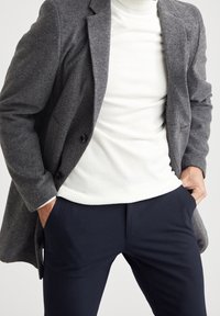 Grå uldfrakke, hvid ribbet turtleneck og marineblå bukser. Frakken har et struktureret design med knapper, hvilket skaber et lagdelt look.