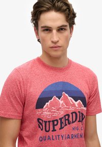 Camiseta de manga corta roja con gráfico de montaña. Texto "SUPERDRY" y "QUALITY GARMENT" en azul oscuro. Tejido de algodón suave y casual.