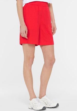 Person, die leuchtend rote Shorts mit elastischem Bund und weiße Sportschuhe trägt, steht mit locker an der Seite hängenden Armen vor einem weißen Hintergrund.