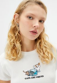 T-shirt blanc à rayures avec un graphique de bande dessinée amusant représentant trois chats et le texte "LES CHATS SONT LIQUIDES" en lettres noires épaisses.