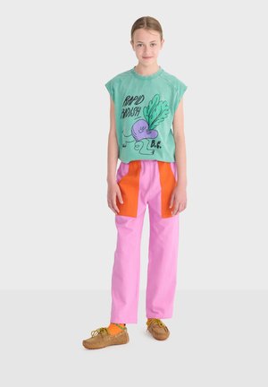 Giovane che indossa una canotta verde con la stampa "Rapid Radish", pantaloni rosa con toppe arancioni, calzini arancioni e mocassini marroni.