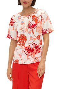 Bloemenblouse in het wit met rode en roze patronen; korte mouwen; ronde halslijn; lichte stof; gecombineerd met felrode broeken.