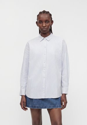 Femme aux cheveux tressés portant une chemise blanche à rayures bleu clair à manches longues et une jupe en jean bleu, debout devant un fond uni.