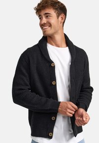 Cárdigan negro de punto con cuello de chal, botones de madera, puños y dobladillo acanalados. Se lleva sobre una camiseta blanca. Tejido texturizado.