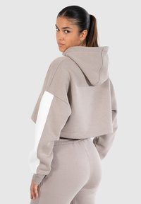 Geknipte, beige hoodie met een grote capuchon en witte accenten op de mouwen, gemaakt van zachte stof met een relaxte pasvorm.