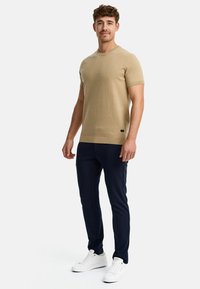 Camisa de punto beige de manga corta con escote redondo, combinada con pantalones azul marino y zapatillas blancas. La camisa presenta puños y dobladillo acanalados.