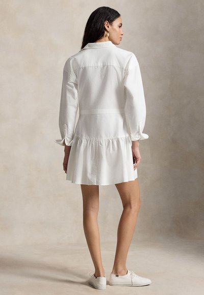 Γυναίκα που φοράει λευκό φόρεμα με μακριά μανίκια και βολάν στη φούστα, καθώς και λευκά δερμάτινα αθλητικά παπούτσια με κορδόνια, στέκεται μπροστά σε μπεζ φόντο.