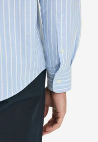 Vaaleansininen raidoitettu kauluspaita, jossa on pitkät hihat nappikäänteillä ja kaareva helma, valmistettu rakenteellisesta kankaasta.