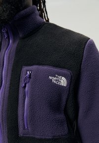 Lähikuvassa musta ja lilla fleece-jakku, jossa on vetoketjullinen rintatasku ja valkoinen "The North Face" -logo vasemmalla puolella.