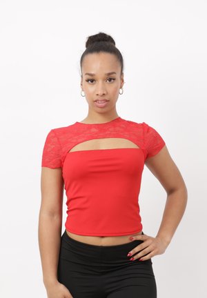 Femme avec les cheveux en chignon, portant un crop top rouge à manches courtes en dentelle et un pantalon noir, posant avec une main sur la hanche.