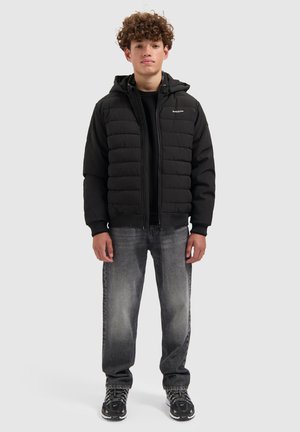 Zwart donzen jack met capuchon, voorzien van doorgestikte delen. Gecombineerd met donkergrijze jeans en zwarte sneakers, effen achtergrond.