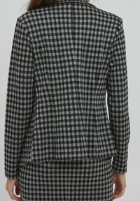 Kobieta w dopasowanej czarno-szarej w kratę marynarce i pasującej spódnicy, widziana z tyłu na jednolitym tle.