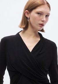 Zwarte wikkeltop van glad materiaal. Heeft een diep V-hals en lange mouwen, met een geplooid ontwerp rond de taille.