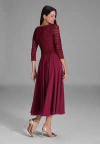 Robe bordeaux avec corsage en dentelle et manches trois-quarts ; jupe fluide ; portée avec des sandales à talons dorés ; texture lisse ; design élégant.