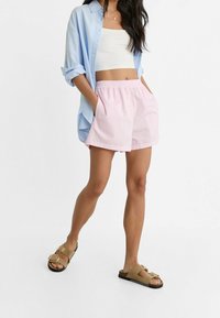 Femme portant une chemise oversize bleu clair, un crop top blanc, un short rose et des sandales à boucle beige, se tenant les mains dans les poches sur fond blanc.