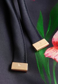 Marine stof met florale patronen, voorzien van gouden hardware labels met de tekst "by Ted Baker." Glad oppervlak en zichtbare stiksels.