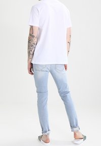 Witte T-shirt met korte mouwen en lichtblauwe jeans met een slim fit, rafelige randen, en witte instap schoenen. Tatoeages zichtbaar op de onderarm.