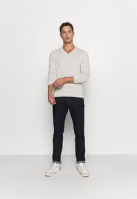 Hellbeige V-Ausschnitt Strickpullover mit langen Ärmeln, kombiniert mit dunklen Jeans und weißen Sneakers. Das Model steht aufrecht vor einem weißen Hintergrund.