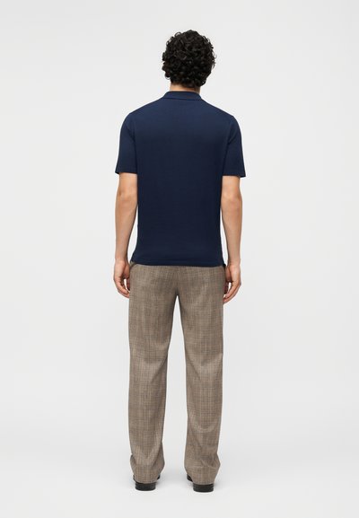 Άνδρας με σγουρά μαλλιά φοράει μπλε σκούρο πόλο με κοντά μανίκια και μπεζ καρό παντελόνια, στέκεται με την πλάτη σε λευκό φόντο.