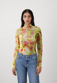 Langærmet gennemsigtig mesh top med et farverigt blomstermotiv i pink og grøn, med høj hals og tætsiddende design. Sammen med blå jeans.