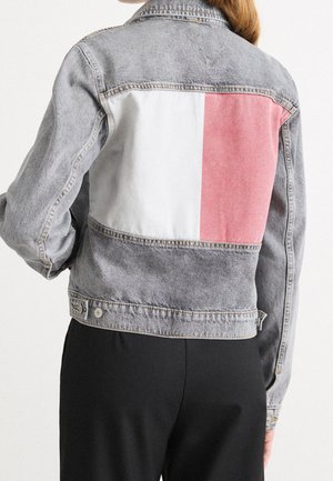 Personne portant une veste en jean grise délavée avec de grands blocs de couleur blancs et roses dans le dos, associée à un pantalon noir.