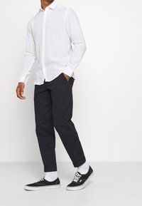 Muž v bílém dlouhém rukávu košili s knoflíky, černých kalhotách s manžetami, bílých ponožkách a černých nízkých teniskách na bílém pozadí.