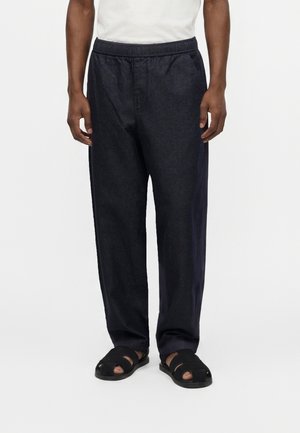 Mann trägt lockere dunkle Hose mit elastischem Bund und schwarze offene Sandalen steht vor schlichtem hellem Hintergrund.