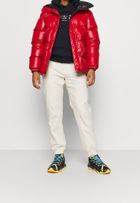 Henkilö, jolla on päällään kiiltävä punainen toppatakki, musta huppari, beige juoksuhousut ja mustat lenkkarit, joissa on siniset nauhat ja keltaiset pohjat.