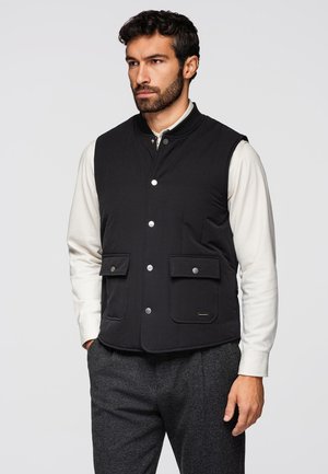 Uomo con barba che indossa un gilet nero abbottonato sopra una camicia bianca a maniche lunghe e pantaloni grigi, in piedi davanti a uno sfondo neutro.